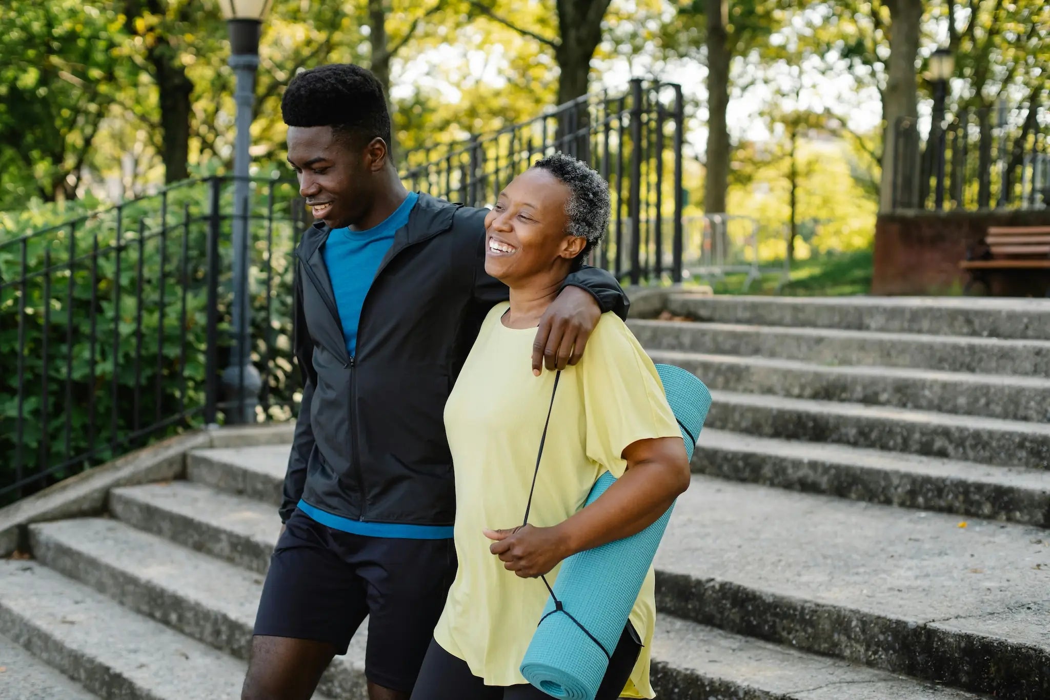 Smiling older woman carrying a yoga mat walking outdoors with a younger man, symbolizing memory support and healthy aging after 50.